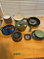 Photo showing assortment of enamel cookware including green pot with lid, blue and yellow pans with wooden handles, brown stoneware pots with lids, and small decorative stoneware dishes.