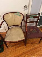 Photo of two antique wooden chairs side by side; one with beige damask upholstery and wooden armrests, the other with floral embroidered seat and wooden slat back.
