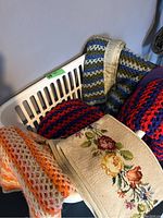 Four colorful crocheted throws and one embroidered wall hanging in a laundry basket and on chair, showing multi-color crochet patterns and floral embroidery detail.