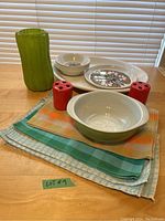 Full lot view showing green Pyrex dish, red toothbrush holders, cloth placemats, floral patterned dishes, and green vase on wooden table.