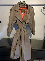 Full view of the taupe vintage London Fog trench coat, showing single button closure, lapels, belt, and adjustable cuff straps.