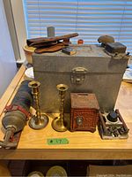 Photo showing various vintage metal novelty and found items including brass candlesticks, a mini safe, Boy Scouts camping utensils in leather case, and a vintage fire extinguisher with pump handle.