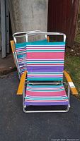 Two aluminum frame folding beach chairs with colorful striped fabric and wooden armrests, standing on pavement outside near a garage area.