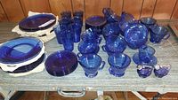 Entire lot spread on table showing multiple cobalt blue glass plates, cups, two-handled soup cups, and salt and pepper shakers.