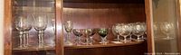 Wide view of multiple crystal stemware pieces in wooden cabinet shelf, showing different styles and some colored glassware in back
