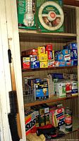 Shelf showing packaged fluorescent bulbs, adapters, multiple boxes of food wraps including Saran Wrap and aluminum foil, and cleaning products such as SOS, Comet, and Sos powder cleansers, plus a flashlight and cloths.