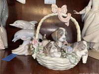 Porcelain figurine of three puppies in woven basket with twisted handle and bow, decorated with pastel flowers. Shelved with other porcelain pieces.