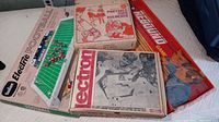Photo of a Tudor Electric Football game board with miniature football players, a red and orange Rebound game box, a Lectron experiment set box, and a portable tape recorder box on a surface.