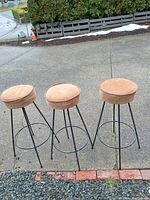 Photo of three vintage bar stools with beige upholstered padded round seats on black angled metal legs arranged outdoors on concrete.