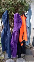 Side view of jackets hanging on a rack outdoors showing navy blue, purple, bright orange, black and light blue jackets arranged in a row.