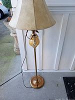 Full view of vintage brass floor lamp standing on floor, attached beige fabric lampshade, brown electrical cord hanging from lamp base.