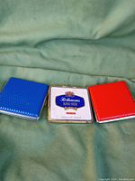 Front view of 3 items: blue and red modern cigarette cases with textured pattern and a vintage Rothmans King Size metal tin with logo on lid.