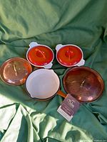 Overview of 2 Descoware mini lidded cast iron pots with orange enamel lids, 1 Palm mini enamelled skillet with white interior and orange handle, 2 copper lids with brass handles.