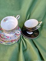 Two stacks of vintage porcelain cups and saucers, one floral set and one black and gold hand-painted set.