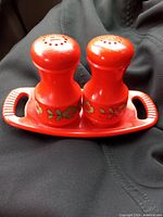 Photo showing pair of red ceramic salt and pepper shakers with green and white floral pattern and matching tray with handles.