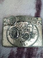 Front view of the metal belt buckle showing embossed John Deere Model D tractor and banner detail.