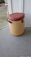 Large insulated soup carrier with beige ribbed plastic body and maroon lid, side handles, and metal latch closure, placed outside near a white wall
