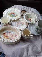 Image of Wedgwood and Royal Doulton china pieces featuring Peter Rabbit and Bunnykins characters. Includes plates, bowls, mugs, and figurines on white cloth.