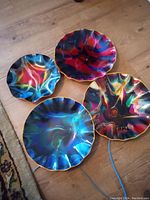 Four vibrant ruffled glass plates in psychedelic swirl colors with gold edges, arranged on wood surface.