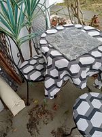 Set shows round table covered with black, white, and gray geometric cloth and a gray marble slab on top, two wrought iron chairs with matching patterned cushions, placed on outdoor concrete patio.