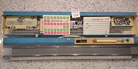 Top-down view of Brother KH588 knitting machine showing the full needle bed, control panel with dials and buttons, and included punch cards and metal tools laid out in the case.