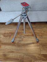 Photo showing the fully extended silver metal professional tripod with three legs and red rubber feet on a wooden floor in front of a couch.