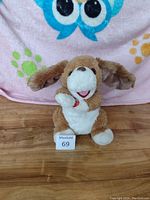 Front view of dancing dog plush toy on wooden floor with colorful blanket background showing details of face, paws, and body.