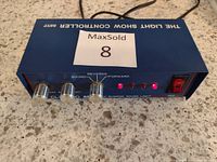 Light show controller blue metal box with control knobs, power switch, and red indicator lights on front panel. Black power cord visible.