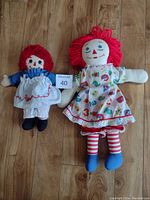 Front view of two Raggedy Ann dolls, 19" and 13", placed on wooden floor showing detailed faces and outfits