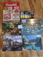 Photo showing all six sealed puzzles including the Campbell's Soup puzzle box set and five individual landscape/cityscape puzzles.