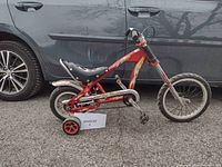 Side view of the vintage Pacific Coast Choppers bicycle showing red frame, black seat, rear training wheel, and front wheel.