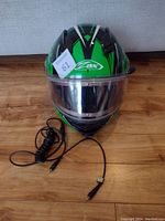 Full front view of ZOX helmet laid on flat surface showing green and black design with clear visor and attached communication cord coiled next to it.