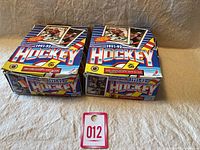 Two unopened boxes of 1991-92 OPC hockey cards placed side by side on a fabric surface with a tag numbered 012 visible