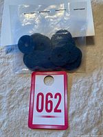 Photo of a small sealed plastic bag containing multiple round dark metal tokens with a center hole and red/white paper tag with number 062.