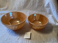 Two vintage Fire King peach orange lustre swirl mixing bowls side by side on cloth surface, large and medium size visible.