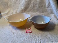Photo showing two Pyrex Cinderella mixing bowls side by side on a fabric backdrop, one in small brown size and one in medium orange size, both with white interiors and pouring spouts.