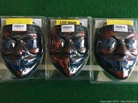 Three LED masks in sealed plastic packaging placed side by side on a green surface, showing front view with colored LED patterns.