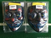 Two packaged LED masks resembling Guy Fawkes masks, showing front with LED light patterns in red and blue under clear plastic packaging.