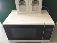 White Hamilton Beach microwave oven with black digital keypad on right side, shown on countertop with two boxes behind it.