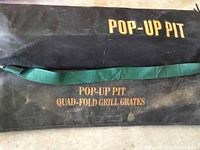 Black carrying case labeled 'Pop-Up Pit Quad-Fold Grill Grates' with green strap lying flat, showing branding and proof of item identity.