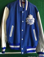 Front view of the Toronto Maple Leafs varsity jacket showing blue wool body, white leather sleeves, front button closure, and embroidered maple leaf logo patch.