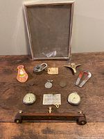 Full lot view showing various items on wooden surface including framed glass, lock, stopwatches, coins, shoehorn, pin, magnet, and control arm.