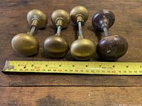 Four door knobs laid out with a measuring tape showing length approximately 3 inches. Three knobs are brass tone finish, in oval shape. Fourth knob is brown tone with surface wear.