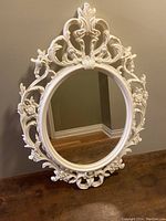 Front view of the oval mirror showing the ornate white frame with floral and scroll decorations on wall background.