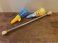 Full view of the metal baton with wooden ends, missing rubber grip, and two plastic toy ball poppers in blue and yellow placed on a wooden surface.