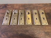 Lot of 6 vintage brass tone door knob plates displayed on wooden surface, showing the full lot and their relative condition and size.