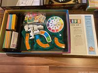The Game of Life board game with components including spinner, curved track pieces, cards, and instructions, all inside the game box.