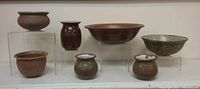 Group photo showing seven ceramic vessels on clear stands including jars, bowls, and a lidded pot.