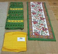 Green and yellow tablecloth folded, floral table runner with green border beside it, and three folded yellow serviettes.