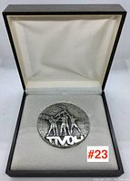 Close view of medallion #23 inside presentation box on white cushioned interior, showing detailed figurative design and partial markings.